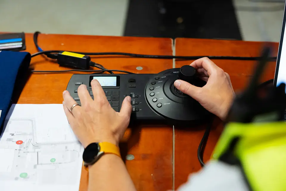 Handen op de camerajoystick naast de plattegrond van het terrein in de controlekamer van Rijvers Festival